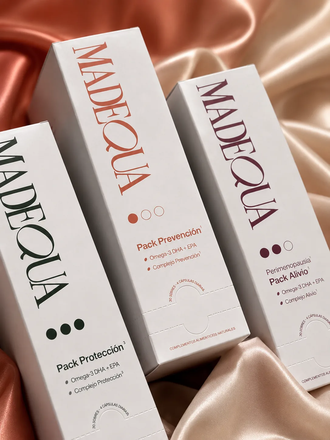 Tres estuches de suplementos Madequa —Pack Protección, Prevención y Alivio— sobre seda, cada uno con un color distintivo